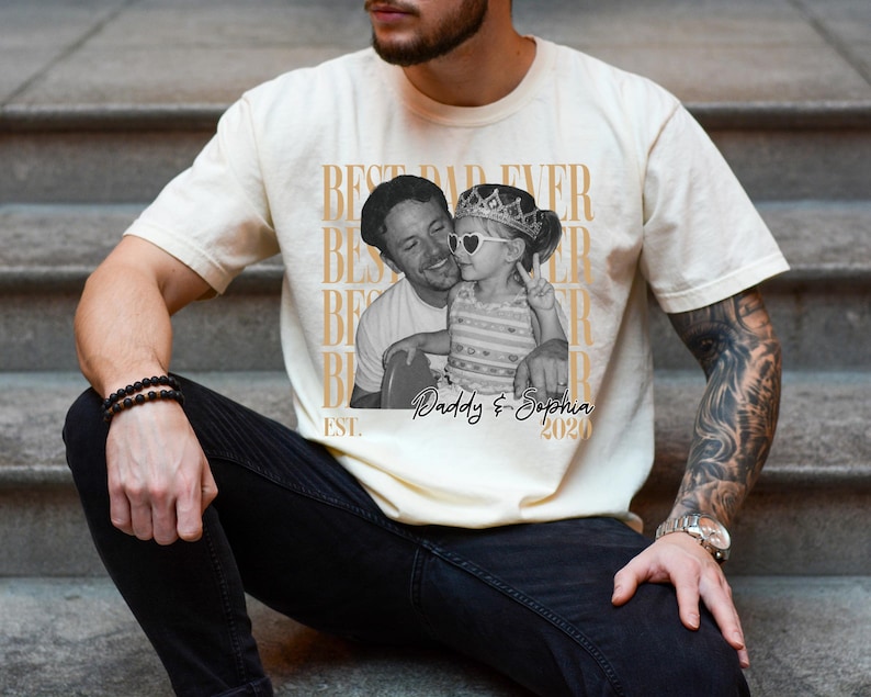 Op de afbeelding: Cr&egrave;mekleurig T-shirt met een zwart-wit foto van een man en een kind. Het shirt heeft de tekst "BEST DAD EVER" verticaal herhaald en "Daddy & Sophia 2020" onder de foto. Het shirt wordt door een persoon gedragen.