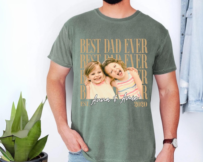 May include: Sage green t-shirt with the text "BEST DAD EVER" in a repeating pattern. The shirt features a photo of two young girls with the names "Anna & Amie" and the year "2020" printed below the photo.