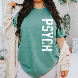 May include: A teal green short-sleeve t-shirt featuring the word PSYCH printed vertically in distressed white capital letters on the front. The shirt has a crew neckline and a relaxed fit, paired with a cream-colored open-front knit cardigan.