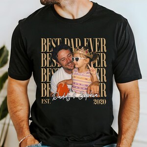 May include: Black t-shirt with a photo of a man and a child, with the text "BEST DAD EVER" repeated vertically in a gold color. The photo shows the man holding a heart-shaped object and the child wearing a crown and heart-shaped sunglasses. The text "Daddy & Sophia" and "2020" are also present.