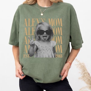 May include: An olive green short-sleeve t-shirt featuring a black and white photograph of a child wearing sunglasses and sticking out their tongue while making a peace sign. The text ALEY'S MOM and EST. 2013 is printed in a tan serif font.
