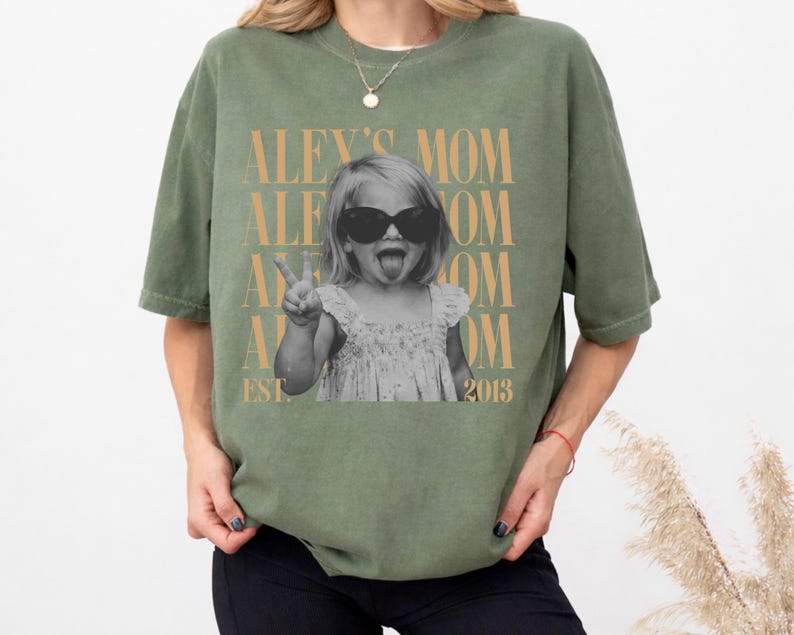 May include: An olive green short-sleeve t-shirt featuring a black and white photograph of a child wearing sunglasses and sticking out their tongue while making a peace sign. The text ALEY'S MOM and EST. 2013 is printed in a tan serif font.