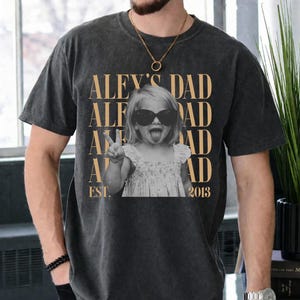 K&ouml;nnte beinhalten: Dunkelgraues T-Shirt mit einem Schwarz-Wei&szlig;-Foto eines Kindes mit Sonnenbrille und Peace-Zeichen. Der goldfarbene Text "ALEX'S DAD" steht &uuml;ber dem Foto, darunter "EST. 2013".