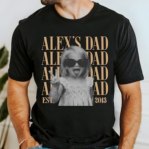 K&ouml;nnte beinhalten: Schwarzes T-Shirt mit einem Schwarz-Wei&szlig;-Foto eines Kindes mit Sonnenbrille, das ein Friedenszeichen macht. Der Text "ALEX'S DAD" ist vertikal in Gold gestapelt, mit "EST. 2013" darunter.