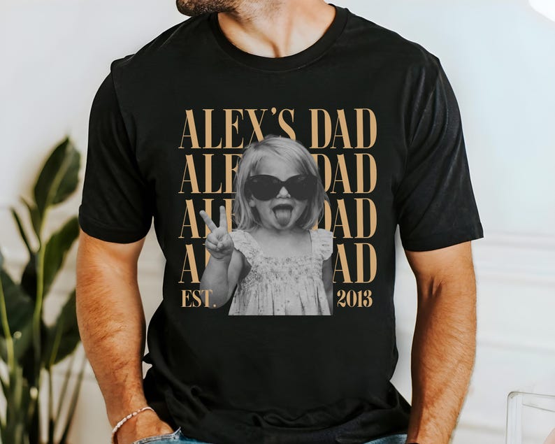 K&ouml;nnte beinhalten: Schwarzes T-Shirt mit einem Schwarz-Wei&szlig;-Foto eines Kindes mit Sonnenbrille, das ein Friedenszeichen macht. Der Text "ALEX'S DAD" ist vertikal in Gold gestapelt, mit "EST. 2013" darunter.