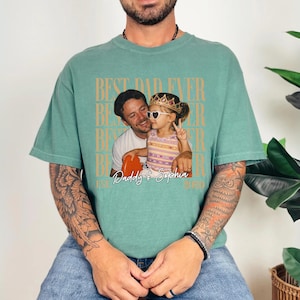 May include: A teal-colored t-shirt with a photo of a man and a child wearing sunglasses and a crown. The shirt has the text "BEST DAD EVER" in a repeating pattern, with "Daddy & Sophia" and the year 2020 below the photo.