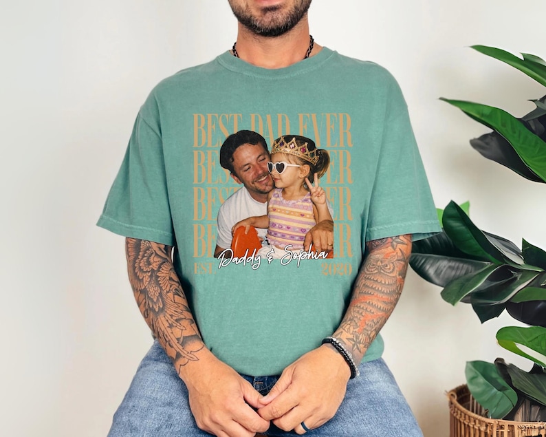 May include: A teal-colored t-shirt with a photo of a man and a child wearing sunglasses and a crown. The shirt has the text "BEST DAD EVER" in a repeating pattern, with "Daddy & Sophia" and the year 2020 below the photo.