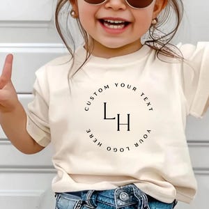 May include: A cream-colored t-shirt with the text "CUSTOM YOUR TEXT HERE YOUR LOGO" encircling the initials "LH" in a circular design. The shirt is worn by a child, who is smiling and wearing sunglasses.