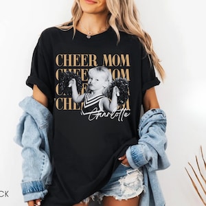 May include: Black t-shirt with the text "CHEER MOM" in gold lettering. A black and white image of a young girl holding pom-poms is centered on the shirt, with the name "Charlotte" in script below. The word "BLACK" is at the bottom left.