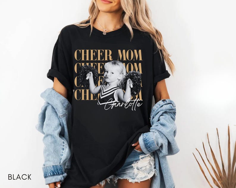 May include: Black t-shirt with the text "CHEER MOM" in gold lettering. A black and white image of a young girl holding pom-poms is centered on the shirt, with the name "Charlotte" in script below. The word "BLACK" is at the bottom left.