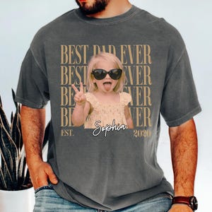 May include: Dark gray t-shirt with the text "BEST DAD EVER" repeated vertically in a tan color. A photo of a young girl wearing sunglasses and sticking her tongue out is in the center. The name "Sophia" and the year "2020" are also printed on the shirt.