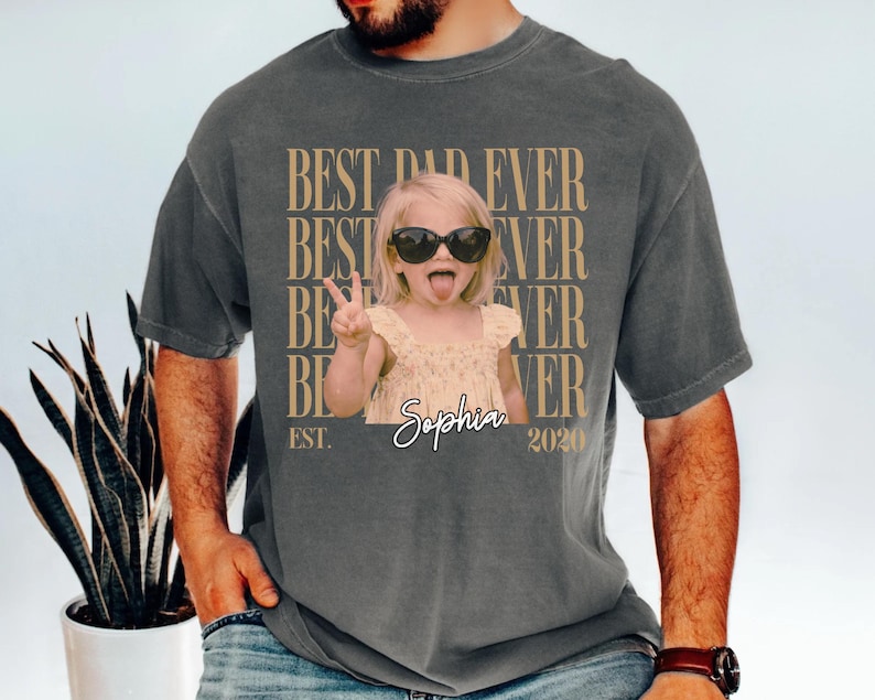 May include: Dark gray t-shirt with the text "BEST DAD EVER" repeated vertically in a tan color. A photo of a young girl wearing sunglasses and sticking her tongue out is in the center. The name "Sophia" and the year "2020" are also printed on the shirt.