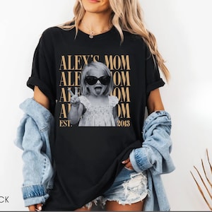 May include: A black short-sleeve t-shirt featuring a graphic print of a child wearing sunglasses and making a peace sign. The text reads ALEY'S MOM repeated in gold lettering with EST. 2013 at the bottom. The shirt is worn with a light blue denim shirt.
