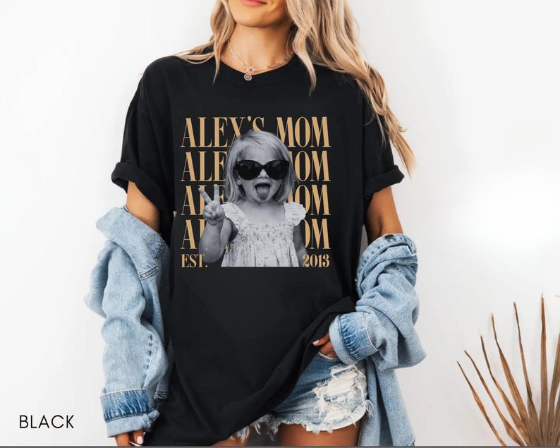 May include: A black short-sleeve t-shirt featuring a graphic print of a child wearing sunglasses and making a peace sign. The text reads ALEY'S MOM repeated in gold lettering with EST. 2013 at the bottom. The shirt is worn with a light blue denim shirt.