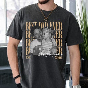 Op de afbeelding: Donkergrijs t-shirt met de tekst "BEST DAD EVER" herhaald. Een zwart-wit foto van een man en kind staat in het midden van het shirt, met de tekst "Daddy & Sophia 2020" eronder. Een casual kledingstuk.
