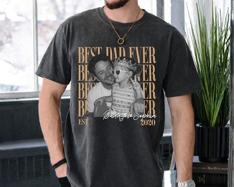 Op de afbeelding: Donkergrijs t-shirt met de tekst "BEST DAD EVER" herhaald. Een zwart-wit foto van een man en kind staat in het midden van het shirt, met de tekst "Daddy & Sophia 2020" eronder. Een casual kledingstuk.