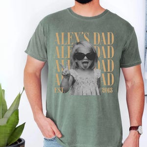 K&ouml;nnte beinhalten: Gr&uuml;nes T-Shirt mit einem Schwarz-Wei&szlig;-Foto eines Kindes mit Sonnenbrille, das ein Friedenszeichen macht. Der Text "ALEX'S DAD" ist vertikal in Gold wiederholt, mit "EST. 2013" darunter.