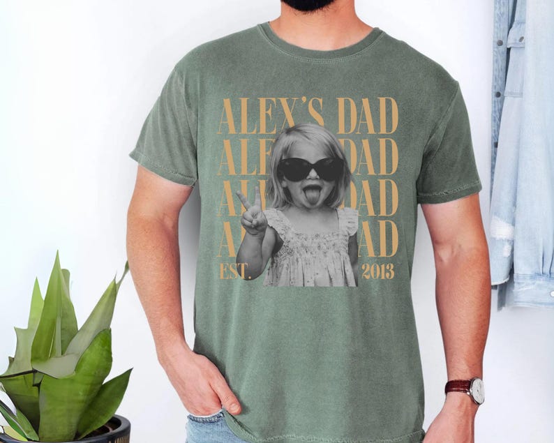 K&ouml;nnte beinhalten: Gr&uuml;nes T-Shirt mit einem Schwarz-Wei&szlig;-Foto eines Kindes mit Sonnenbrille, das ein Friedenszeichen macht. Der Text "ALEX'S DAD" ist vertikal in Gold wiederholt, mit "EST. 2013" darunter.