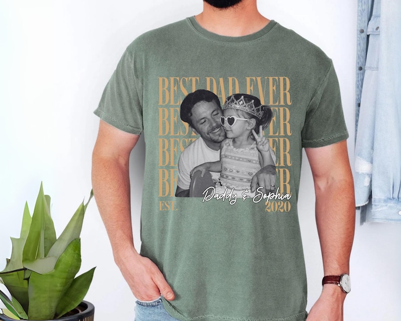 Op de afbeelding: Saliegroen t-shirt met een zwart-wit foto van een vader en kind. De tekst "BEST DAD EVER" is goudkleurig en verticaal herhaald. Daaronder staat "Daddy & Sophia 2020".