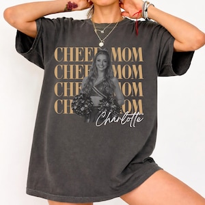 May include: Dark gray t-shirt with the words "CHEE MOM" repeated four times in a gold color. A photo of a cheerleader is in the center, with the name "Charlotte" in a script font below.