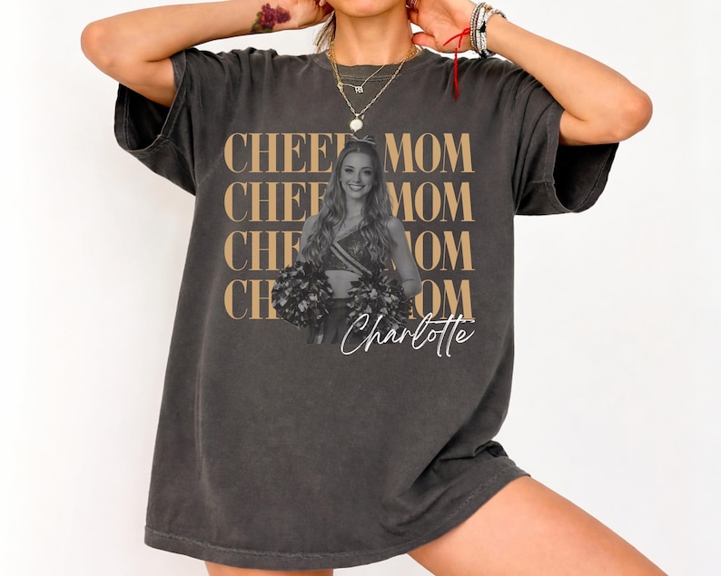 May include: Dark gray t-shirt with the words "CHEE MOM" repeated four times in a gold color. A photo of a cheerleader is in the center, with the name "Charlotte" in a script font below.