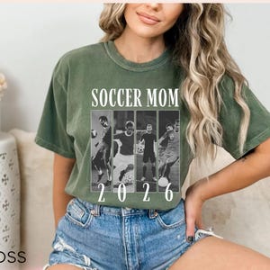 May include: Olive green t-shirt with the text "SOCCER MOM" above a black and white photo collage of soccer players. The year "2026" is printed below the collage. The model is wearing blue denim shorts.