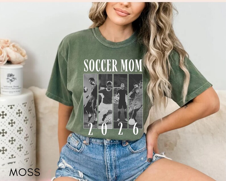 May include: Olive green t-shirt with the text "SOCCER MOM" above a black and white photo collage of soccer players. The year "2026" is printed below the collage. The model is wearing blue denim shorts.