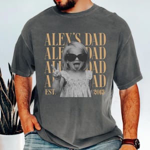 K&ouml;nnte beinhalten: Dunkelgraues T-Shirt mit einem Aufdruck, der ein Schwarz-Wei&szlig;-Foto eines Kindes mit Sonnenbrille und Peace-Zeichen zeigt. Der Text "ALEX'S DAD" ist vertikal in Gold wiederholt, darunter "EST. 2013".