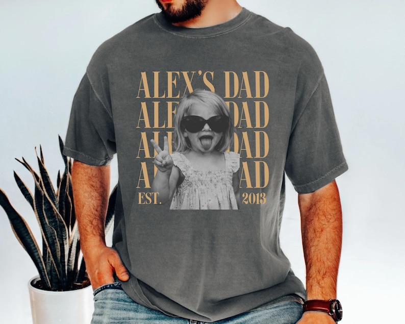 K&ouml;nnte beinhalten: Dunkelgraues T-Shirt mit einem Aufdruck, der ein Schwarz-Wei&szlig;-Foto eines Kindes mit Sonnenbrille und Peace-Zeichen zeigt. Der Text "ALEX'S DAD" ist vertikal in Gold wiederholt, darunter "EST. 2013".