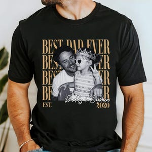 Op de afbeelding: Zwart t-shirt met een sepia-kleurige afbeelding van een man en een kind. Het kind draagt een kroon en hartvormige zonnebrillen. De tekst op het shirt luidt "BEST DAD EVER" en "Daddy & Sophia 2020".