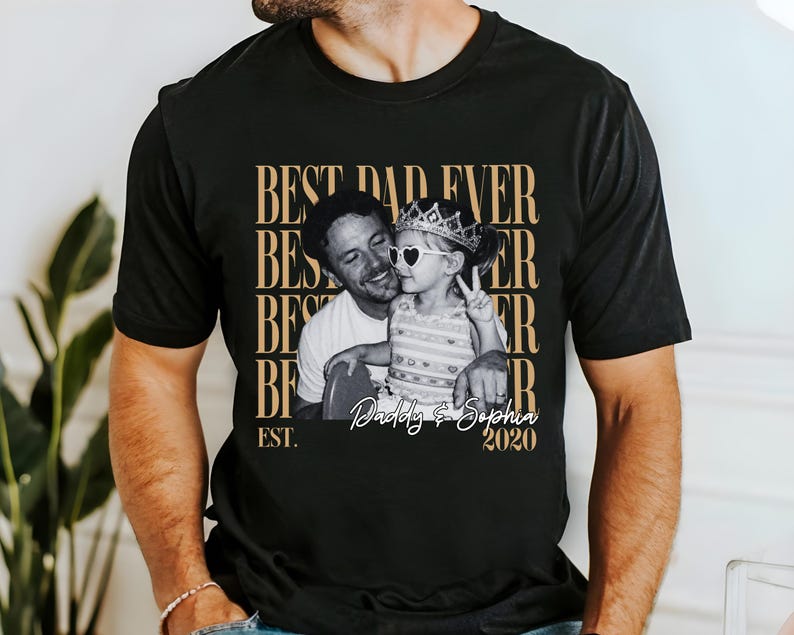 Op de afbeelding: Zwart t-shirt met een sepia-kleurige afbeelding van een man en een kind. Het kind draagt een kroon en hartvormige zonnebrillen. De tekst op het shirt luidt "BEST DAD EVER" en "Daddy & Sophia 2020".