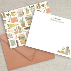 May include: A stationery set featuring a peach-colored envelope and two note cards. One card displays a repeating pattern of books, plants, lamps, and pennants with the text READ MORE. The other card is white with the text A NOTE FROM Allison Keller.