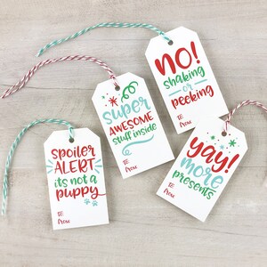 May include: Four white gift tags with colorful twine. The tags have different humorous sayings, including "Spoiler Alert, it's not a puppy", "Super Awesome Stuff Inside", "No! Shaking or Peeking", and "Yay! More Presents".