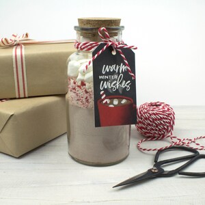 May include: A glass jar filled with hot chocolate mix, marshmallows, and crushed peppermint, topped with a cork and a tag that reads "Warm Winter Wishes." The jar is tied with red and white twine, and is next to wrapped gifts.