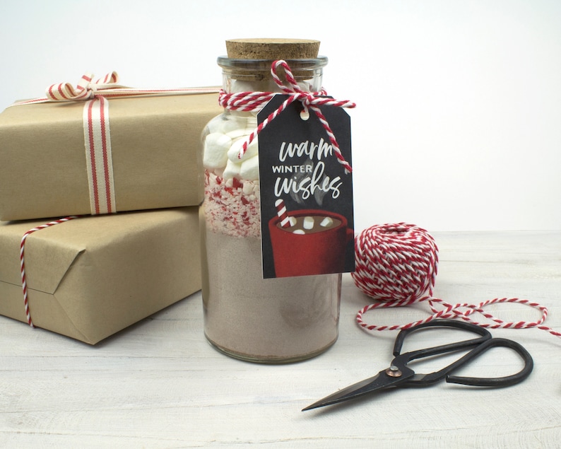 May include: A glass jar filled with hot chocolate mix, marshmallows, and crushed peppermint, topped with a cork and a tag that reads "Warm Winter Wishes." The jar is tied with red and white twine, and is next to wrapped gifts.