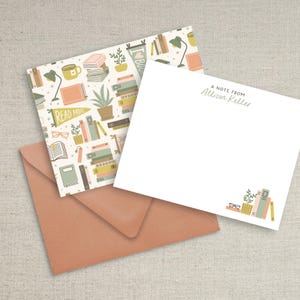 May include: A set of personalized stationery featuring a white card with the text "A note from Allison Keller" and a patterned envelope with a design of books, plants, and glasses. The envelope has a light pink background with a white pattern.