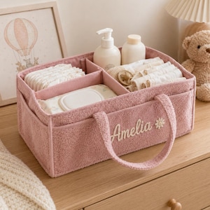 May include: A dusty pink textured fabric nursery organizer with two handles, featuring the embroidered name Amelia and a small daisy icon on the front. The interior is divided into three sections containing baby diapers, wipes, bottles, and a pacifier.