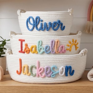 Peut inclure: Trois paniers en corde blanche empil&eacute;s sur une &eacute;tag&egrave;re en bois, chacun avec le nom d'un enfant en &eacute;criture color&eacute;e : Oliver, Isabella et Jackson. Un &eacute;l&eacute;phant en bois et une petite plante en pot sont &eacute;galement sur l'&eacute;tag&egrave;re. Une d&eacute;coration murale arc-en-ciel est en arri&egrave;re-plan.