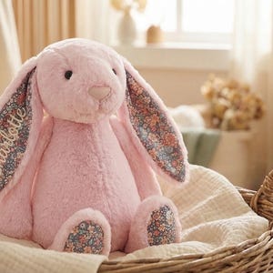 May include: A plush pink bunny rabbit with long ears and floral patterned fabric inside the ears and on the feet. The name "Charlotte" is embroidered on one ear. The bunny is seated in a woven basket.