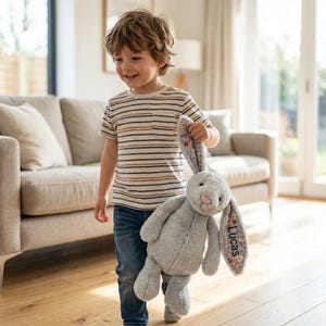 May include: A gray plush bunny with patterned inner ears and the name "Lucas" embroidered on one ear. The bunny is held by a child wearing a striped t-shirt and jeans. The background includes a sofa and wooden floor.