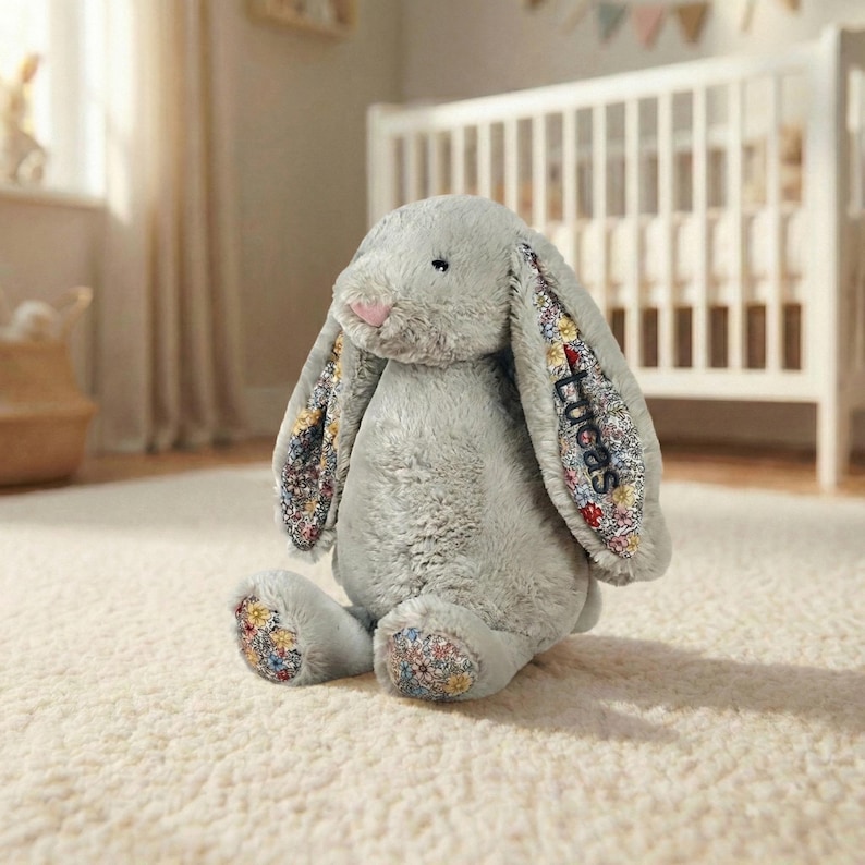 May include: A plush gray bunny rabbit with long floppy ears. The inside of the ears and the soles of the feet are decorated with a colorful floral pattern. The name "Lucas" is embroidered on one ear. The bunny has a pink nose.