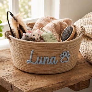 Peut inclure: Un panier en corde beige rempli de jouets et d'accessoires pour animaux de compagnie. Le panier porte le nom "Luna" en bleu et une empreinte de patte. Il contient des souris en peluche, une brosse et un jouet &agrave; plumes. Pos&eacute; sur une surface en bois.