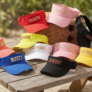 Könnte beinhalten: Eine Auswahl an Sonnenblenden in verschiedenen Farben, darunter Rot, Rosa, Gelb, Blau und Schwarz, jede mit einem anderen Namen bestickt. Die Sonnenblenden sind auf einem hölzernen Picknicktisch mit Sonnenbrille und Getränk ausgestellt.