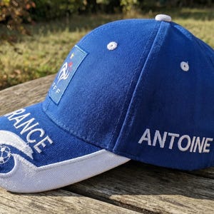 May include: A blue baseball cap with white accents and the word "FRANCE" embroidered on the brim. The cap features the French flag and the name "ANTOINE" embroidered on the side. A white button is on top.