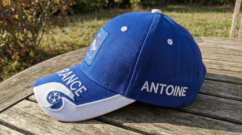May include: A blue baseball cap with white accents and the word "FRANCE" embroidered on the brim. The cap features the French flag and the name "ANTOINE" embroidered on the side. A white button is on top.