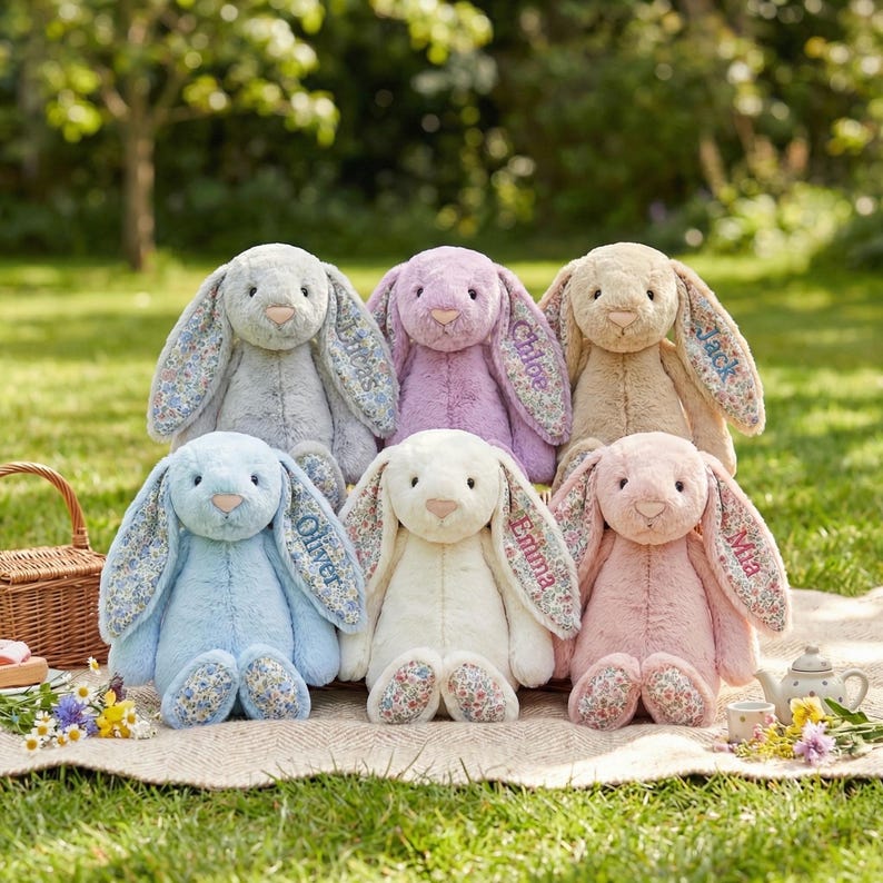 May include: Six plush bunny rabbits in various pastel colors, including blue, pink, and lavender, are arranged on a blanket. Each bunny has floral patterned ears and feet, with a name embroidered on one ear. A wicker basket and tea set are in the background.