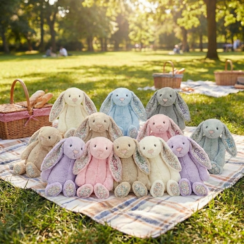 May include: A collection of plush bunny rabbits in various pastel colors, including pink, lavender, and blue. Each bunny has long ears with floral patterns and embroidered names. The bunnies are arranged on a plaid blanket outdoors.