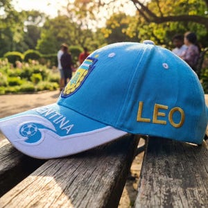 May include: A light blue baseball cap with the word "LEO" embroidered in gold on the side. The cap features the Argentina national team logo and the word "ARGENTINA" on the brim. The cap is resting on a wooden bench.