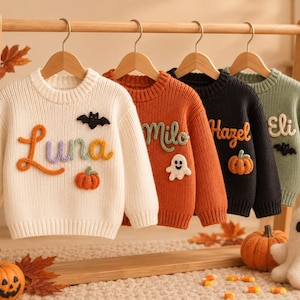 May include: Four long-sleeve knit sweaters in cream, orange, black, and sage green hanging on wooden hangers. Each sweater features a name embroidered in cursive yarn, including Luna, Milo, Hazel, and Eli, with small felted Halloween motifs.