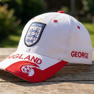May include: White baseball cap with red accents and the word "ENGLAND" on the brim. Features the England national team emblem and the name "GEORGE" embroidered in red. A soccer ball graphic is on the brim.